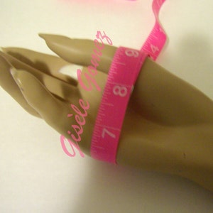 May include: A pink measuring tape wrapped around a mannequin hand. The tape shows measurements in inches, with the 7-inch mark visible. The background is a plain white surface. The image has the text "Gisele Gomez" in pink.