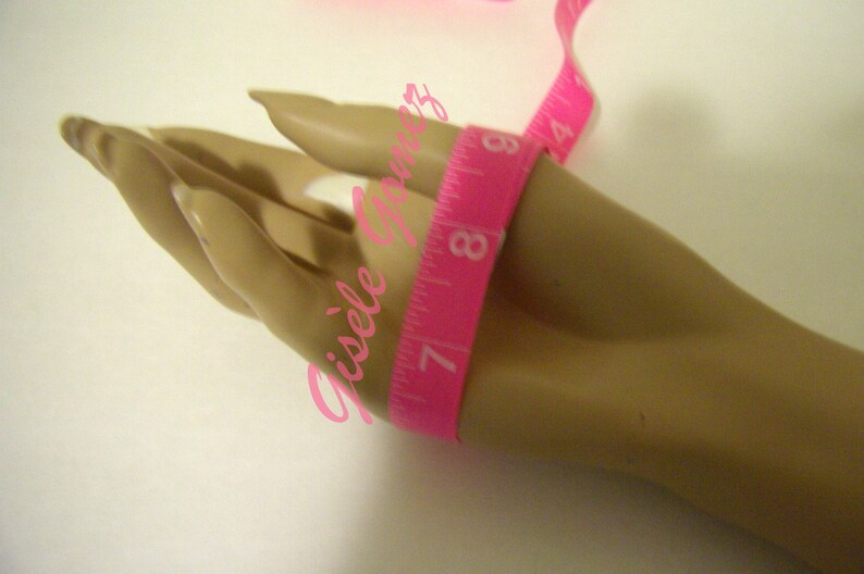 May include: A pink measuring tape wrapped around a mannequin hand. The tape shows measurements in inches, with the 7-inch mark visible. The background is a plain white surface. The image has the text "Gisele Gomez" in pink.