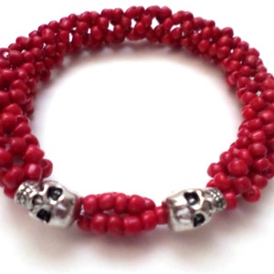May include: A red beaded bracelet with two silver skull accents. The bracelet is made of small, round red beads woven together. The skulls are detailed with eye sockets and a nose cavity.
