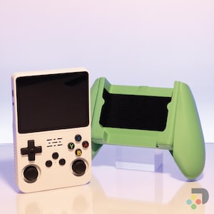 Puede incluir: Una consola de juegos portátil blanca con un mando verde. La consola tiene una pantalla negra, un pad direccional y botones etiquetados como A, B, X e Y. El mando tiene una empuñadura verde y una pantalla negra.