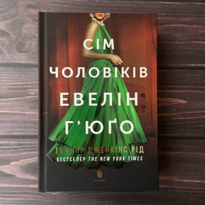 以下が含まれることがあります： 緑のドレスを着た女性が表紙の書籍。タイトルは「エブリン・フーゴの七人の夫」テイラー・ジェンキンス・リード著。ニューヨーク・タイムズ紙のベストセラー