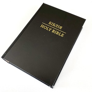 Puede incluir: Una Biblia de tapa dura negra con letras doradas. El texto en la portada dice "БІБЛІЯ HOLY BIBLE". El libro está cerrado e inclinado, sobre un fondo blanco.