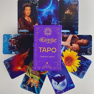 Könnte beinhalten: Ein violettes Tarotkartenspiel mit den Worten "Giggle TARO" in goldener Schrift. Das Deck ist von Tarotkarten mit verschiedenen Illustrationen umgeben, darunter eine Frau, eine schwarze Katze und eine Sonnenblume. Die Karten sind nummeriert.