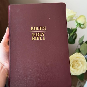 Puede incluir: Una Biblia encuadernada en cuero burdeos con letras doradas. El texto en la portada dice "БІБЛІЯ HOLY BIBLE". El libro está sostenido en una mano, con un ramo de rosas blancas en el fondo.