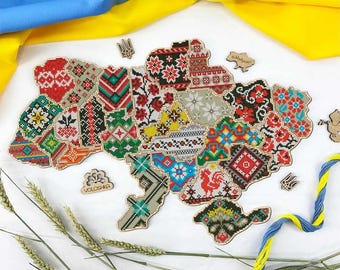 May include: A wooden map of Ukraine with a colorful embroidered design. The map is made up of individual pieces that fit together. The map is surrounded by wheat stalks and a blue and yellow ribbon.
