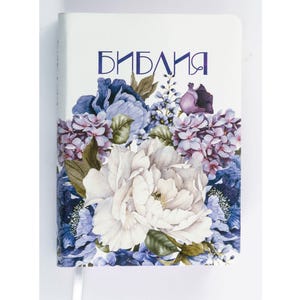 Könnte beinhalten: Ein Hardcover-Buch mit dem Wort "БИБЛИЯ" in blauer Schrift. Das Cover zeigt ein Aquarell-Blumenmuster mit weißen, lila und blauen Blumen. Ein weißes Leseband ist sichtbar.