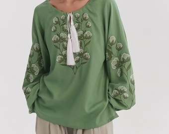 May include: A green blouse with long sleeves, adorned with floral embroidery in green and white. It has a round neckline with a tassel tie. The blouse is worn with beige trousers. The embroidery is in shades of green and white.