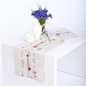 May include: A beige linen table runner featuring embroidered vertical floral patterns in red, blue, yellow, and green. A white ceramic teapot and matching teacup sit on the runner, accompanied by a glass vase filled with blue cornflowers.