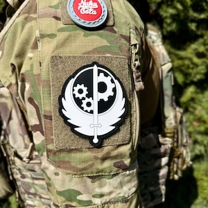 Può includere: Una toppa bianca e nera con una spada e ingranaggi, con il testo "Nuka Cola" su una toppa rossa e bianca su una giacca mimetica.