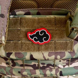 May include: A black cloud-shaped patch with white swirls and a red outline is affixed to a camouflage tactical vest. The patch is centered on a brown velcro panel, with the vest featuring various straps and mesh.