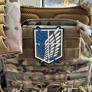 May include: A patch with a stylized emblem of blue and white wings on a grey background, attached to a camouflage tactical vest. The patch is rectangular with a black border. The vest has multiple pockets and straps.
