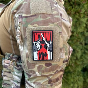 Puede incluir: Un parche rojo con la palabra "KYIV" en letras blancas. El parche presenta una ilustración en blanco y negro de un grupo de personas que sostienen armas y están de pie frente a un fondo rojo.