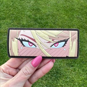 May include: A rectangular patch with anime-style eyes and heart-shaped pupils. The eyes are blue with black eyeliner and long lashes. The hair is blonde, and the skin is pink with blush. The patch has a black border.