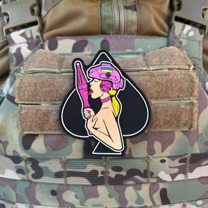 Può includere: Una toppa nera a forma di picche presenta una donna dei cartoni animati con un casco rosa che tiene in mano un oggetto rosa. La donna ha i capelli biondi e tira fuori la lingua. La toppa è su uno sfondo mimetico.