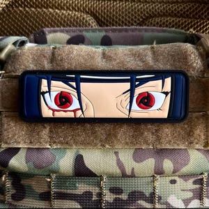 May include: A rectangular patch depicting anime-style eyes with red irises and black outlines. The eyes have a teardrop design and are set against a tan skin tone. The patch has a blue border and is set against a camouflage background.