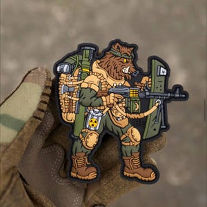 May include: A green, brown, and black embroidered patch depicting a cartoon bear-like creature wearing a military uniform and carrying a machine gun. The creature is wearing a helmet and has a backpack with a radiation symbol on it. The patch has the text "BE" on it.