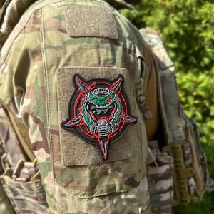 May include: A black, red, and green patch featuring a stylized skull design with a pentagram border. The patch is affixed to a camouflage tactical vest. The image is taken outdoors with green foliage in the background.
