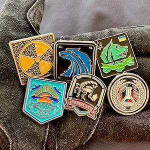 May include: A selection of enamel pins with diverse designs. The pins showcase a yellow radiation symbol, a blue eagle, a green wolf, a turquoise shield with a sunrise, a scorpion, and a science emblem. Each pin has a metal border.