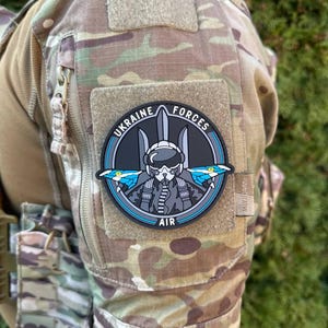 Könnte beinhalten: Ein schwarzer und grauer Patch mit einem blauen Umriss. Der Patch zeigt ein stilisiertes Bild eines Piloten mit Helm und Sauerstoffmaske, über dem sich das ukrainische Dreizack-Symbol befindet. Der Text "UKRAINE FORCES AIR" ist um das Bild herumgedruckt.