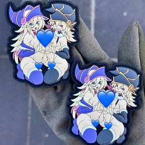 Może przedstawiać: Dwa haftowane naszywki przedstawiające postać z anime w kreskówkowym stylu z blond włosami, ubraną w fioletowo-czarny kapelusz czarownicy i niebieską sukienkę. Postać trzyma w każdej ręce niebieskie serce. Naszywki znajdują się na ciemnozielonym tle z tkaniny.