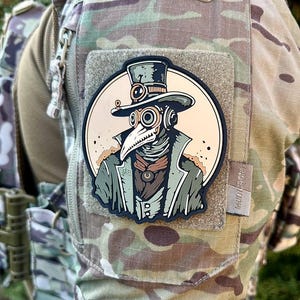 Puede incluir: Un parche en blanco y negro con una ilustración estilizada de una persona que lleva un sombrero de copa, gafas y una máscara de médico de la peste. El parche está sujeto a un uniforme militar de camuflaje.