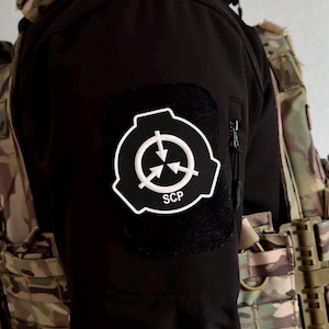 Op de afbeelding: Zwarte jas met een wit en zwart SCP-patch. De patch heeft een cirkelvormig ontwerp met pijlen en de letters "SCP". De jas heeft een zak met ritssluiting en wordt gedragen met een camouflagevest.