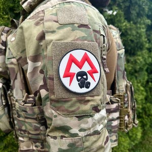 Könnte beinhalten: Taktische Tarnweste mit einem runden Aufnäher mit einem roten Blitzdesign und einem schwarzen Gasmaskensymbol. Die Weste hat mehrere Taschen und einen Klettverschluss zum Anbringen des Emblems. Der Hintergrund ist eine verschwommene Ansicht von grünem Laub.