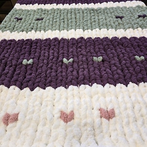 Puede incluir: Una manta de punto grueso con rayas horizontales en blanco, morado oscuro y verde salvia. La manta est&aacute; decorada con peque&ntilde;os detalles en relieve con forma de coraz&oacute;n en colores contrastantes, incluyendo rosa, blanco y verde claro.