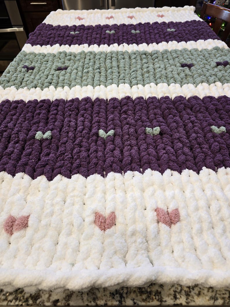 Puede incluir: Una manta de punto grueso con rayas horizontales en blanco, morado oscuro y verde salvia. La manta est&aacute; decorada con peque&ntilde;os detalles en relieve con forma de coraz&oacute;n en colores contrastantes, incluyendo rosa, blanco y verde claro.