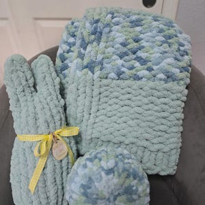 Puede incluir: Un conjunto de crochet en verde menta, azul y blanco que incluye un conejito, una manta y un cojín redondo. El conejito está atado con una cinta de cuadros amarilla. La manta y el cojín tienen una textura de punto grueso.
