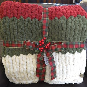 Puede incluir: Una manta de punto grueso con franjas horizontales de colores en rojo oscuro, verde oliva y crema. La manta est&aacute; envuelta con una cinta de cuadros rojos y verdes atada con un lazo, decorada con bayas de acebo y hojas artificiales.