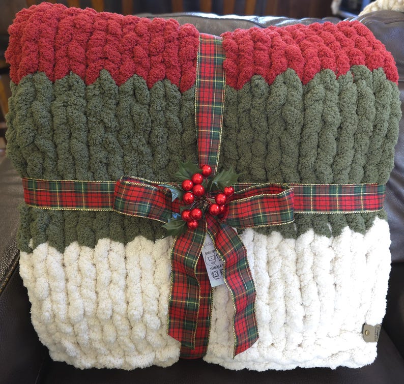 Puede incluir: Una manta de punto grueso con franjas horizontales de colores en rojo oscuro, verde oliva y crema. La manta est&aacute; envuelta con una cinta de cuadros rojos y verdes atada con un lazo, decorada con bayas de acebo y hojas artificiales.