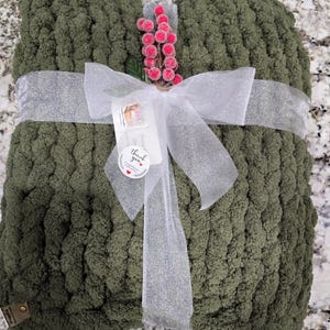Puede incluir: Una manta de punto grueso color verde oliva doblada y atada con una cinta blanca brillante. Una ramita decorativa de bayas rosas est&aacute; insertada en el lazo, junto con una etiqueta blanca que dice thank you y un peque&ntilde;o coraz&oacute;n rojo.