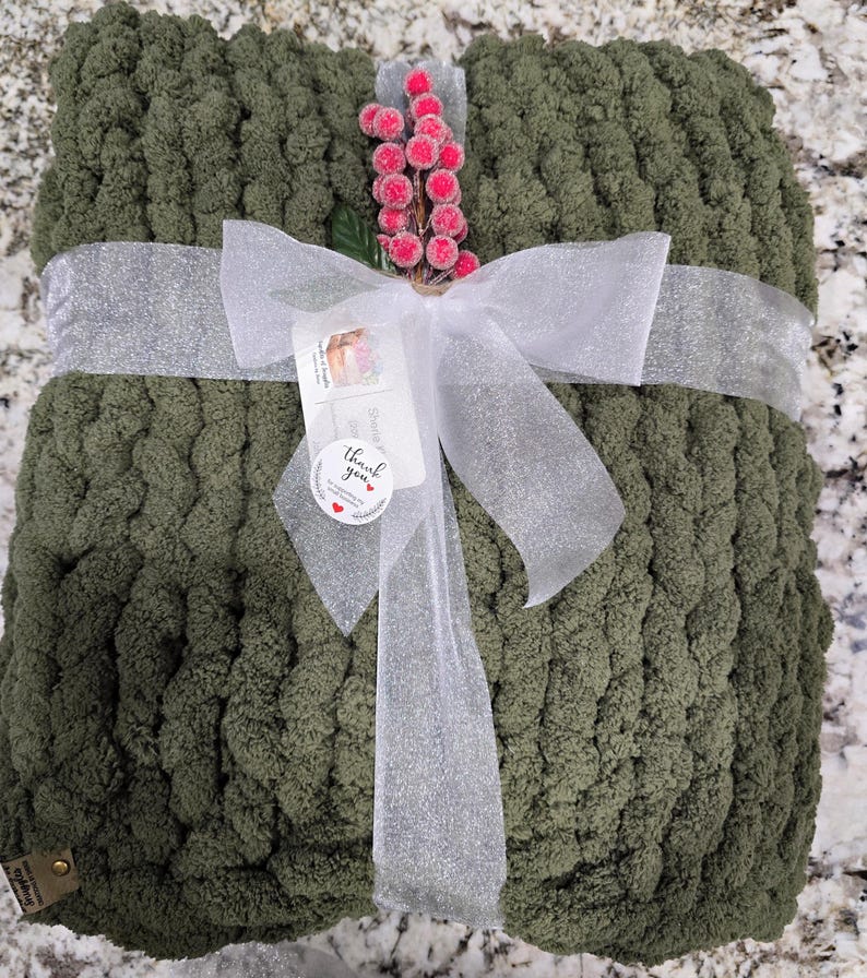 Puede incluir: Una manta de punto grueso color verde oliva doblada y atada con una cinta blanca brillante. Una ramita decorativa de bayas rosas est&aacute; insertada en el lazo, junto con una etiqueta blanca que dice thank you y un peque&ntilde;o coraz&oacute;n rojo.