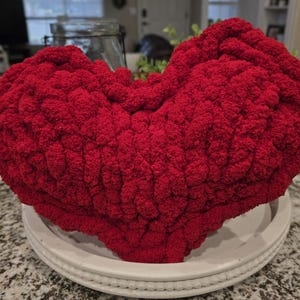 Puede incluir: Un gran cojín decorativo en forma de corazón, de color rojo vibrante. El cojín está hecho de una tela de punto gruesa y texturizada y descansa sobre una bandeja decorativa redonda blanca. Una pieza de acento acogedora y llamativa.