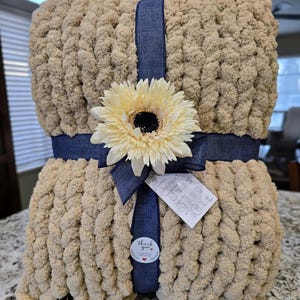 Puede incluir: Una manta de punto grueso en color beige claro, envuelta con una cinta de tela azul oscuro. Un girasol artificial grande de color crema est&aacute; sujeto al centro de la cinta. Se incluye una peque&ntilde;a etiqueta blanca con instrucciones de cuidado.