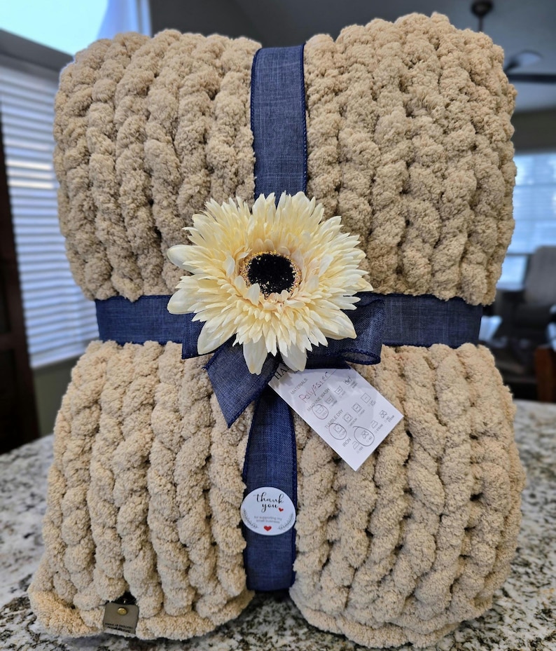 Puede incluir: Una manta de punto grueso en color beige claro, envuelta con una cinta de tela azul oscuro. Un girasol artificial grande de color crema est&aacute; sujeto al centro de la cinta. Se incluye una peque&ntilde;a etiqueta blanca con instrucciones de cuidado.