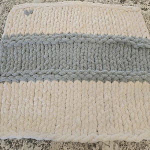 Puede incluir: Una funda de coj&iacute;n cuadrada tejida a mano con lana gruesa. El dise&ntilde;o presenta rayas horizontales en color crema y gris, con un peque&ntilde;o detalle en forma de coraz&oacute;n gris situado en la esquina superior izquierda de la secci&oacute;n crema.