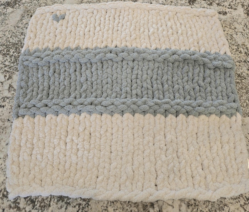 Puede incluir: Una funda de coj&iacute;n cuadrada tejida a mano con lana gruesa. El dise&ntilde;o presenta rayas horizontales en color crema y gris, con un peque&ntilde;o detalle en forma de coraz&oacute;n gris situado en la esquina superior izquierda de la secci&oacute;n crema.