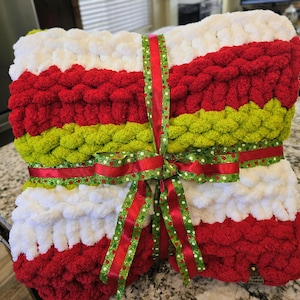Puede incluir: Una manta de punto grueso con rayas horizontales en blanco, rojo y verde lima. La manta est&aacute; doblada y atada con una cinta decorativa roja y verde con un patr&oacute;n de lunares blancos. Tiene una peque&ntilde;a etiqueta rectangular adjunta.