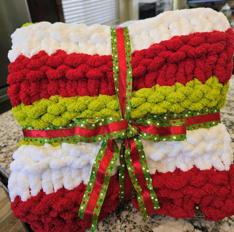 Puede incluir: Una manta de punto grueso con rayas horizontales en blanco, rojo y verde lima. La manta est&aacute; doblada y atada con una cinta decorativa roja y verde con un patr&oacute;n de lunares blancos. Tiene una peque&ntilde;a etiqueta rectangular adjunta.