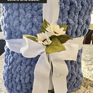 Puede incluir: Una manta de punto grueso azul envuelta en una cinta blanca con dos flores artificiales blancas y hojas verdes. La manta presenta el texto Available in additional colors and sizes! y una peque&ntilde;a etiqueta con thank you.