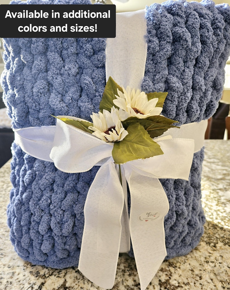 Puede incluir: Una manta de punto grueso azul envuelta en una cinta blanca con dos flores artificiales blancas y hojas verdes. La manta presenta el texto Available in additional colors and sizes! y una peque&ntilde;a etiqueta con thank you.
