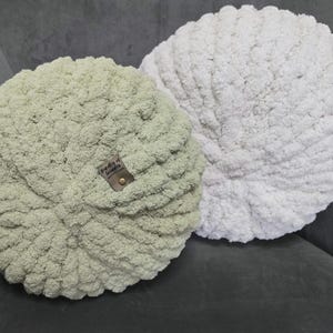 Puede incluir: Dos cojines redondos de punto grueso. Uno es de color verde salvia suave, el otro gris claro. El cojín verde tiene una pequeña etiqueta que dice "freddy of snuggle". Los cojines están sobre un sofá gris oscuro.