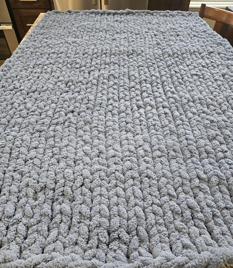 Puede incluir: Una manta rectangular tejida a mano con hilo gris grueso y voluminoso. El tejido presenta un patr&oacute;n de punto de bucle repetitivo y consistente en toda la superficie, creando una apariencia densa, texturizada y suave en toda la tela.