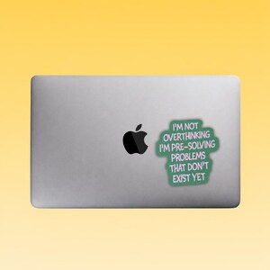 May include: A silver laptop with a black Apple logo. A green sticker with white text that reads "I'M NOT OVERTHINKING I'M PRE-SOLVING PROBLEMS THAT DON'T EXIST YET" is stuck on the laptop's lid.