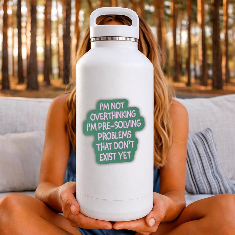 May include: White insulated water bottle with a silver handle and a green sticker that reads "I'M NOT OVERTHINKING I'M PRE-SOLVING PROBLEMS THAT DON'T EXIST YET". The bottle is held by a person.