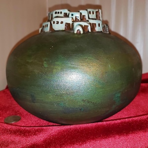 May include: A green ceramic vase with a raised design of a pueblo-style building on top. The vase has a glossy finish and a rounded shape.