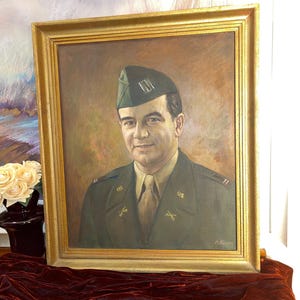 May include: A framed oil painting of a man in military uniform. The portrait features a man wearing a green cap and olive-green uniform with gold insignia. The painting is set against a neutral background, within a gold frame.