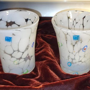May include: Two white glass vases with a unique design. The vases have a flared shape and are decorated with black, gray, and colorful accents. Each vase has a blue label with the word "AMICA" on it. The vases are on a dark red velvet surface.